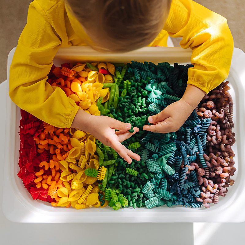 Child leaning over bin of multicolored pasta