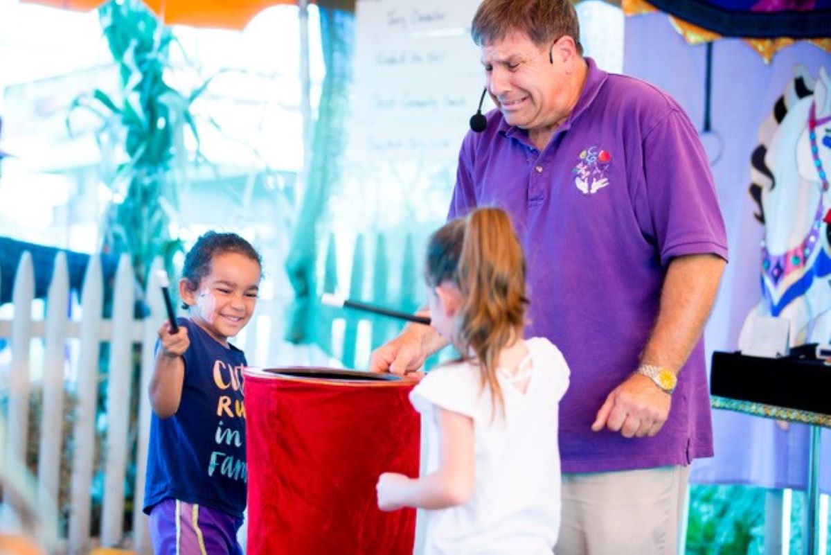 Magician with two children with magic wands surrounding a red bag. 