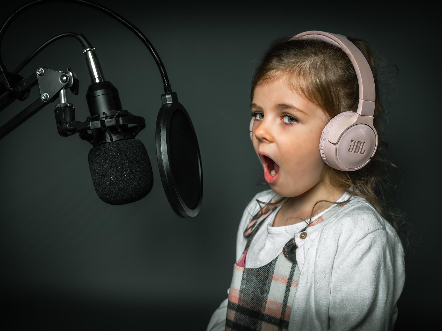 Little girl in white with pink headphones on singing into a microphone.