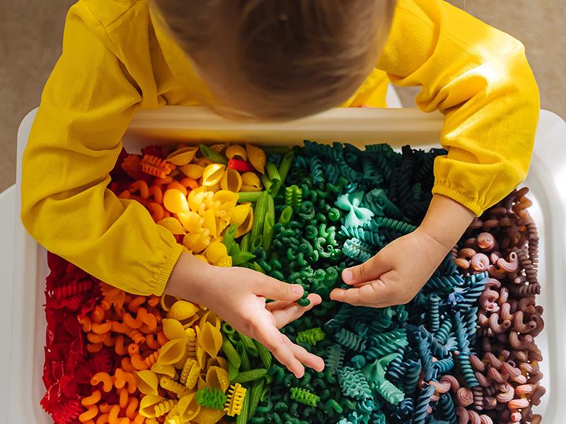 Child leaning over bin of multicolored pasta